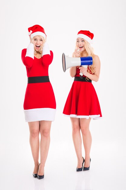 Two playful blonde sisters twins in dresses of santa claus having fun with megaphone over white background