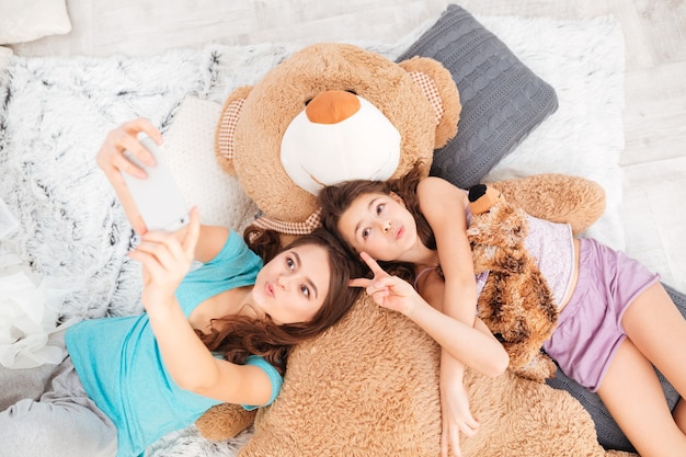 Two playful cute sisters lying, sending kisses and making selfie using smartphone at home