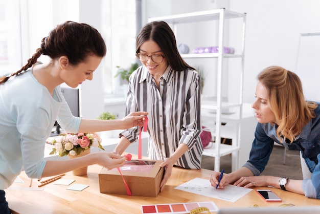 Two pleasant happy tailors tying up a box with a dress with a pink ribbon while their colleague filling out a form