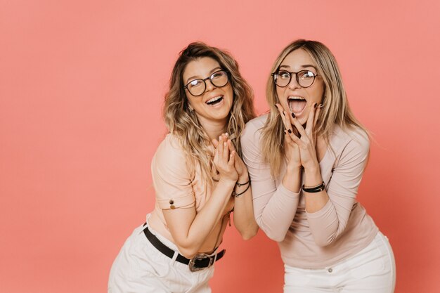 Two pretty blondes in glasses wearing light blouses and white jeans screaming and laughing with wide open mouth, shocked by the surprises they saw. Parties and celebrate, enjoy your life.