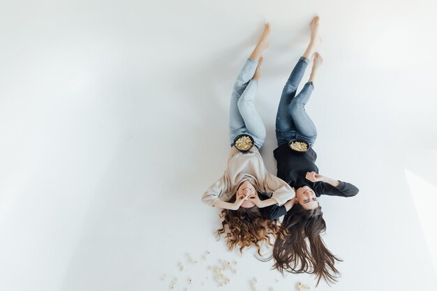 Two pretty cheerful young girls friends standing isolated over white background, eating popcorn