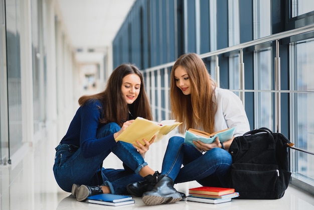 Two pretty female students with books sitting on the floor in the university hallway