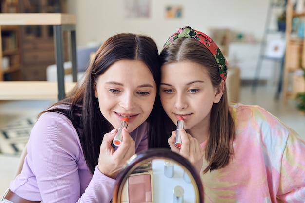 Photo two pretty females with long hair looking in mirror and putting pink and red lipstick on lips after applying nude makeup on their faces