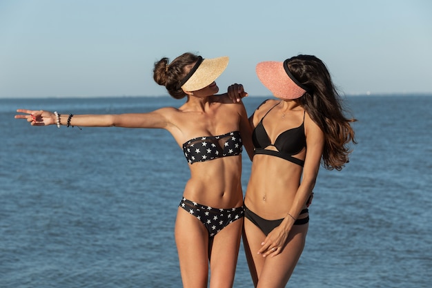 Two pretty slim darkhaired girls in swimsuits and caps stand near the sea on a sunny day