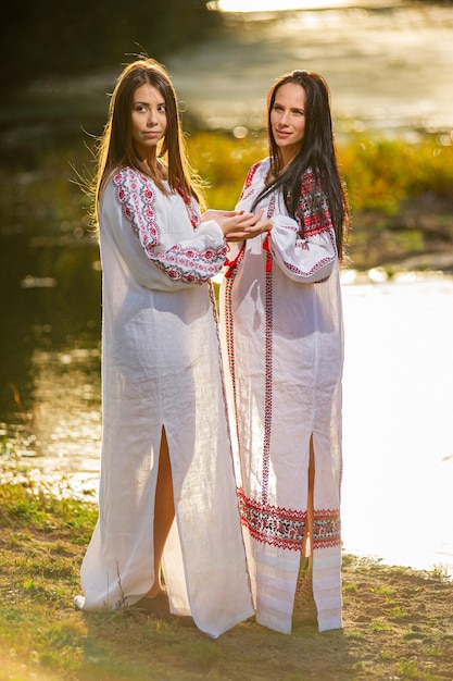 Two Pretty young ladies wear traditional Ukrainian clothes and flower wreath walk in wheat field beautiful ethnic girl in handmade decorated floral crown admire nature Sunset