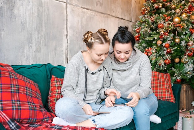 Two pretty young sisters in gray sweatshirts and blue jeans sit on couch near