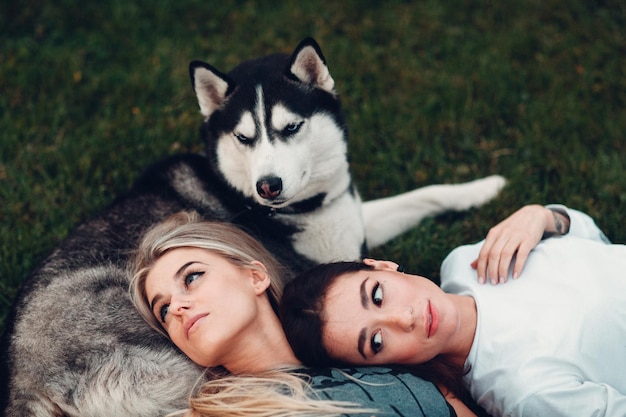 Two pretty young woman laying with husky dog on the grass