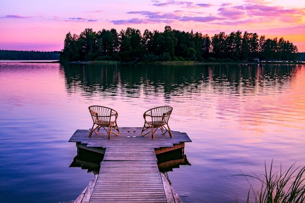 Two rattan wooden chairs and glasses of red wine on a pier overlooking a lake at sunset in Finland