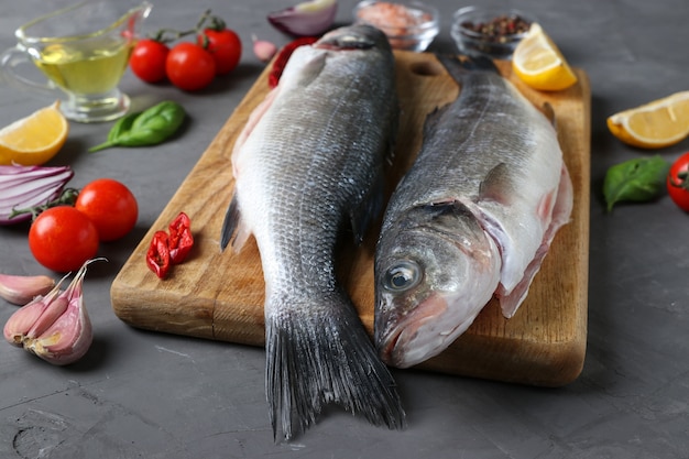 Two raw seabass fishes with ingredients and seasonings like basil, lemon, salt, pepper, cherry tomatoes and garlic on wooden board