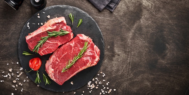 Two raw steak New York  with rosemary and spices on a piece of parchment paper on old dark stone background. Top view. Mock up.