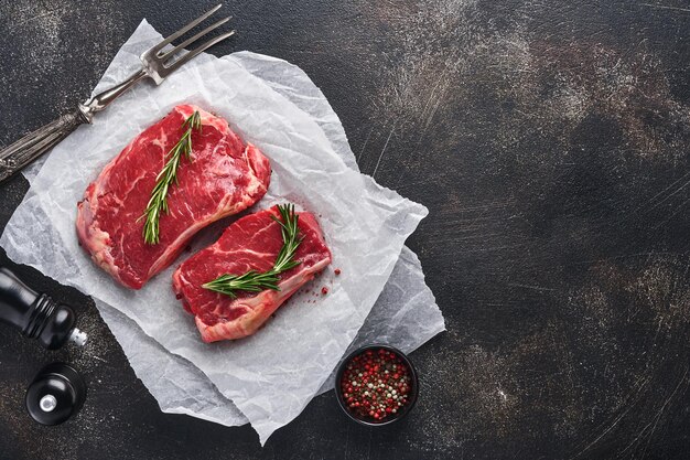 Two raw steak New York with rosemary and spices on a piece of parchment paper on old dark stone background. Top view. Mock up.