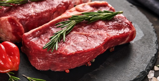 Two raw steak New York with rosemary and spices on a piece of parchment paper on old dark stone background. Top view. Mock up.