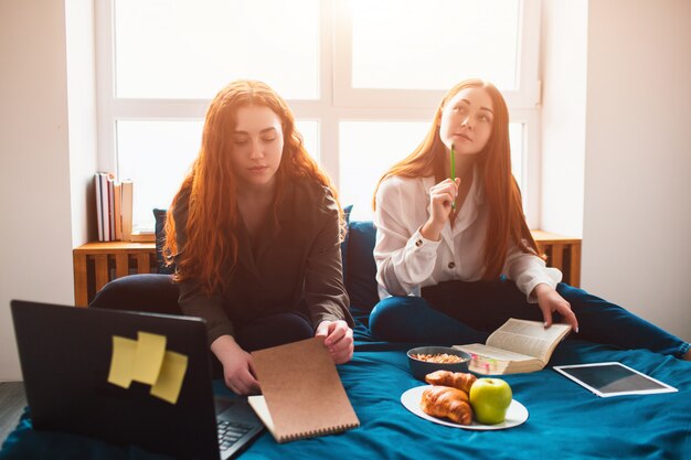 Two red-haired students study at home or prepare for exams. Young women doing homework in a dormitory bed near the window. There are notebooks, food books, a tablet and laptop and documents