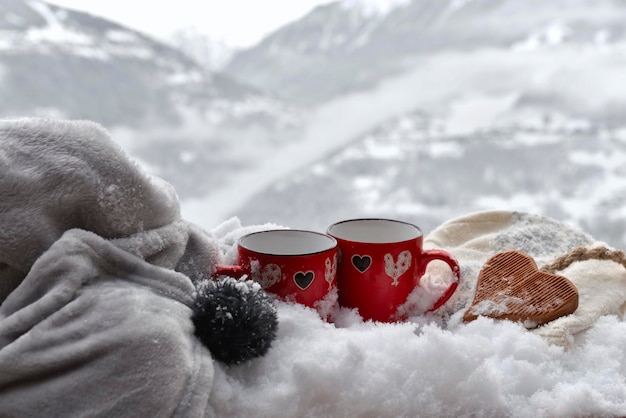 Two red mugs withwooden heart shaped on the snow with blanket on mountain