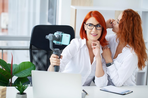 Two redhaired businesswomen in the office smile and do selfie on a smartphone