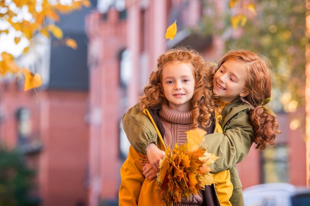 Two redhaired girls sisters cheerfully walk along the city street during the golden autumn leaf fall