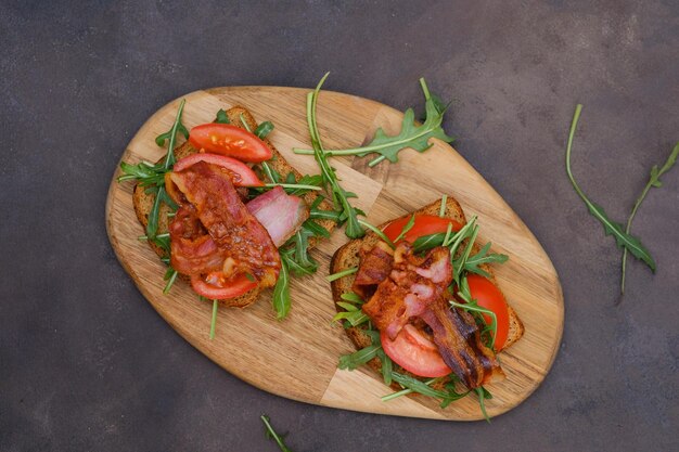 Two sandwiches with aragula fried ham tomatoes on cutboard on dark brown background top view