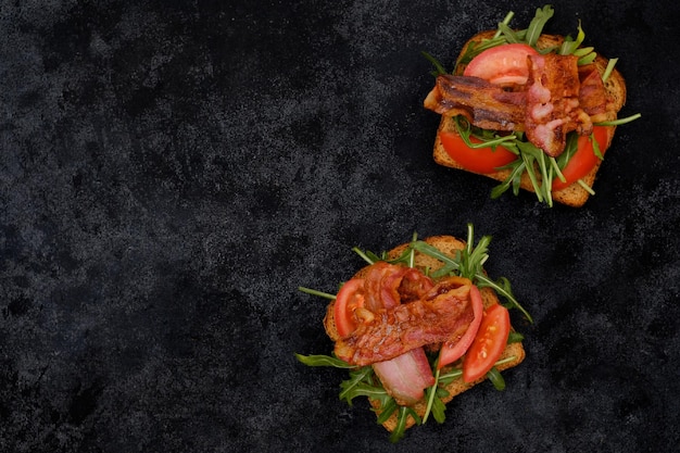 Two sandwiches with aragula fried ham tomatoes on dark background top view