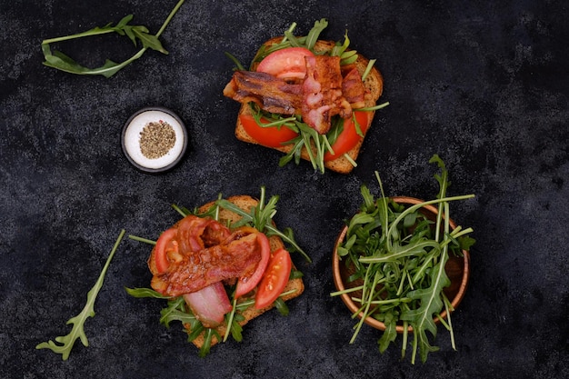 Two sandwiches with aragula fried ham tomatoes on dark background top view