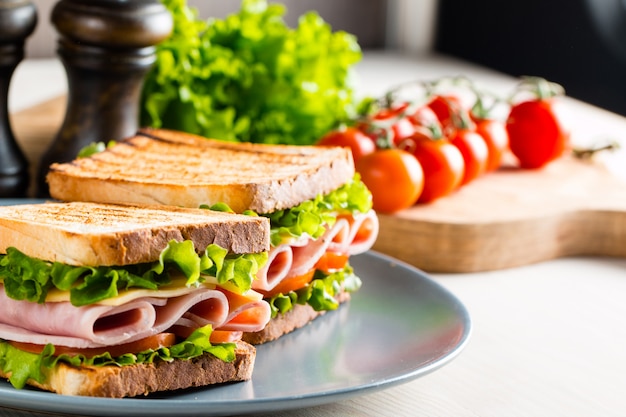 Two sandwiches with ham and fresh vegetables on rustic wooden cutting board.