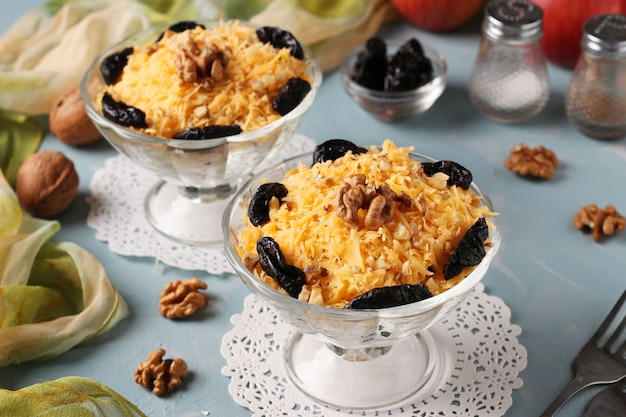 Two servings of salad with chicken, cheese and apples, decorated prunes and walnuts in glass bowls on light blue table, Closeup