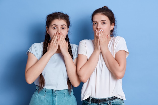 Two shocked women dress whit casual t shirts posing against blue wall covering their mouths