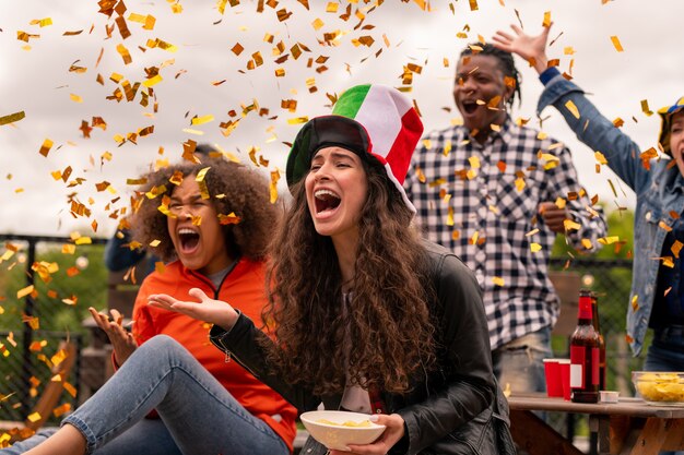 Two shouting female football fans and their friends expressing gladness while throwing confetti during match broadcast