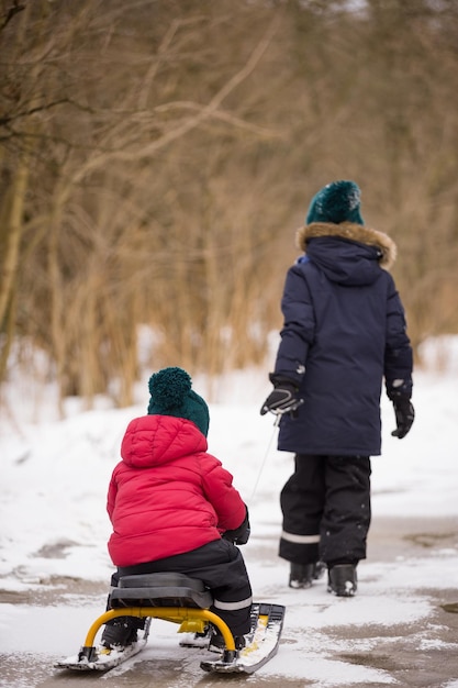 Two siblings walking in a winter park Children outdoors Brother pulling sled with a toddler boy