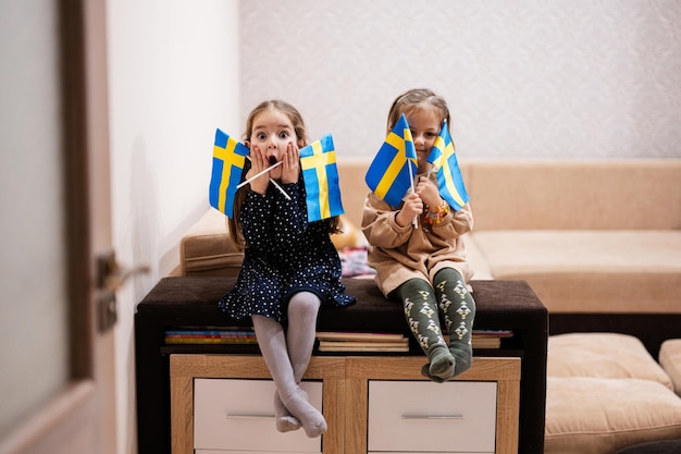 Two sisters are sitting on a couch at home with swedish flags on hands Sweden children girls with flag