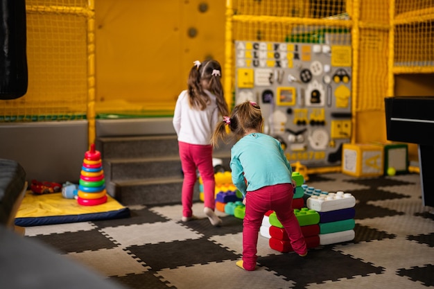 Two sisters playing at kids play center while build with colored plastic blocks