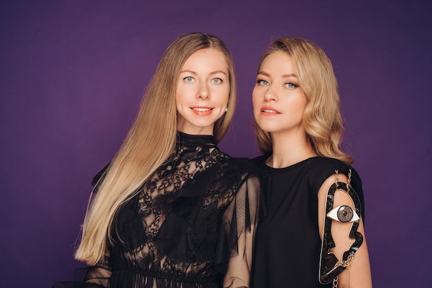 Two sisters pose in the studio on a purple background 2 beautiful women in black fancy dresses gathered for a party