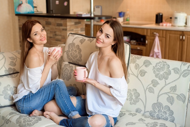 Two sisters on the sofa with a cups of tea in hands. Two best friends enjoying time.