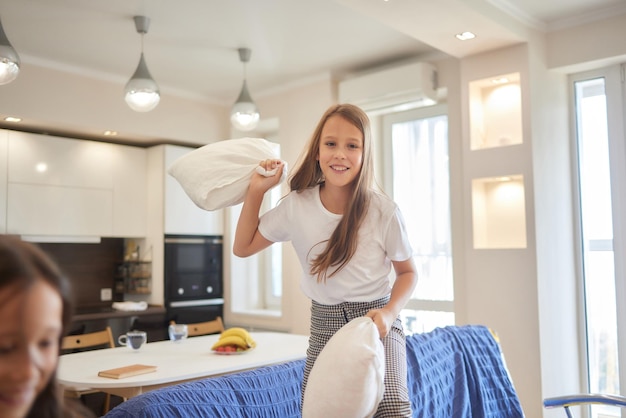 Two sisters wearing white pajamas fight with pillows and have fun concept of youth wellness and heal