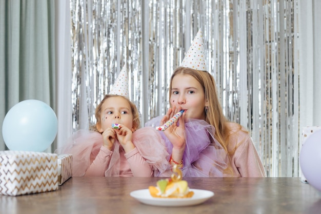 Two sisters of and years old in party hats and elegant dresses are sitting at table and blowing ho