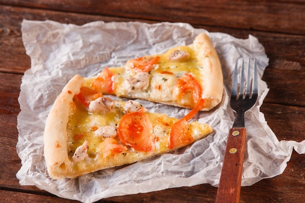 Two slices of fresh baked pizza on white paper napkin with fork, close up view. Italian food, fast lunch served on wooden rustic dark table.