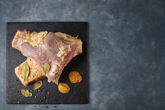 Two slices of raw lard with a layer of meat, with mustard, pepper and bay leaf, against a dark background