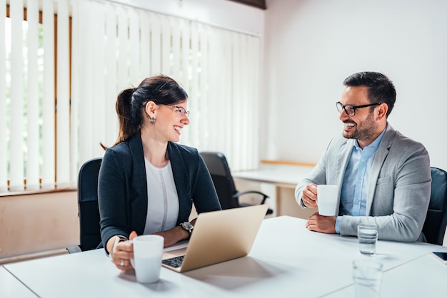 Two smiling business people drinking tea or coffee while sitting at office desk.