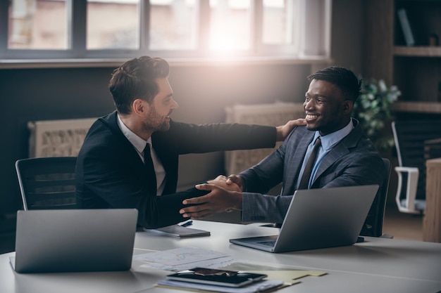 Two smiling businessmen in formalwear shaking hands while sitting at the desk in modern office