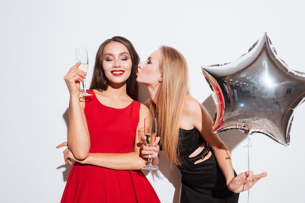 Two smiling cute young women drinking champagne and kissing over white background