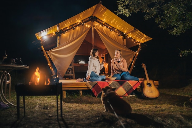 Two Smiling female friends drinking wine and eating fruits sitting in cozy glamping tent in autumn evening bonfire Luxury camping tent for outdoor holiday and vacation Lifestyle concept