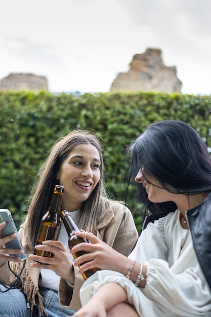 Two smiling female friends toasting with beer sitting on the grass Lifestyle of young female friends sitting on the grass toasting with beer bottles