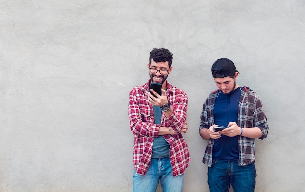 Two smiling friends leaning on a wall checking their cell phones Friends leaning on a wall texting on their phones Friend showing cell phone to his friend Smiling friends checking cell phones