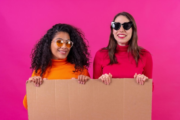 Two smiling multiethnic female friends holding a cardboard sign over a pink background