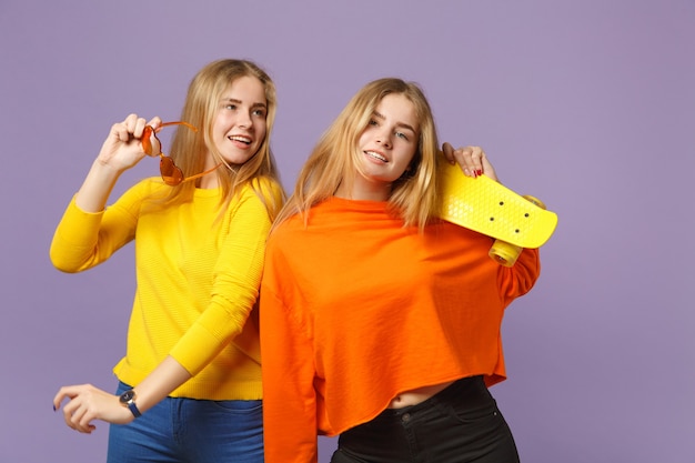 Two smiling young blonde twins sisters girls in vivid clothes heart eyeglasses holding yellow skateboard isolated on pastel violet blue wall. People family lifestyle concept. 