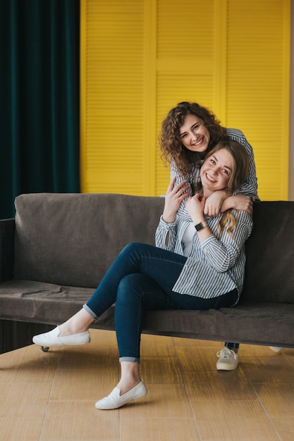 Two smiling young girls in striped shirts, jeans and sneakers posing on the couch. studio shooting.