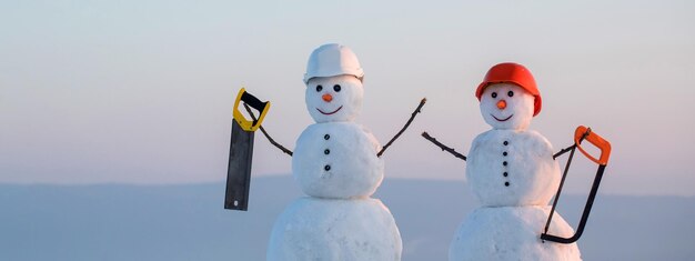 Photo two snowmen builders in building helmet hold saw snowman in hard hat on the snow outdoor background