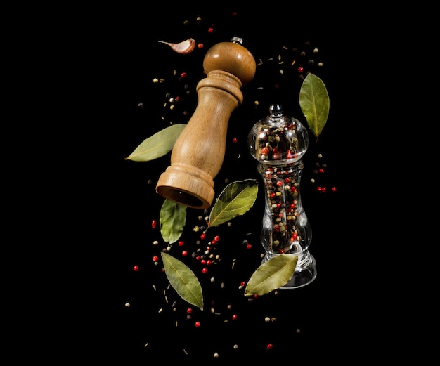 Two spice mills with peppercorns and laurel leaves flying on black