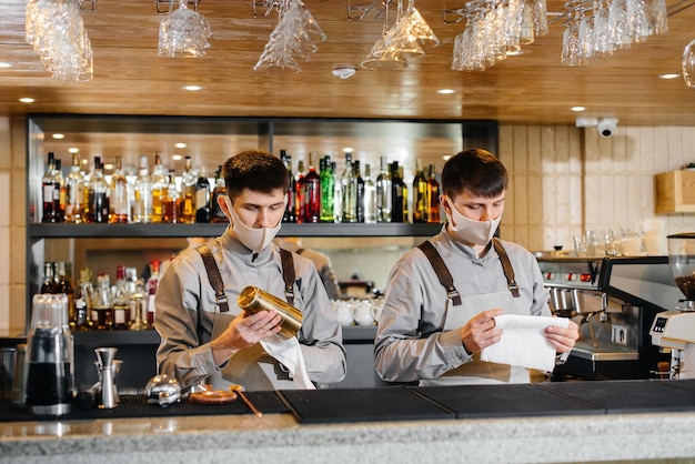 Two stylish bartenders in masks and uniforms during the pandemic, rub glasses to shine. The work of restaurants and cafes during the pandemic.