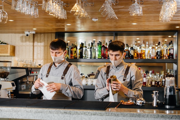 Two stylish bartenders in masks and uniforms during the pandemic, rub glasses to shine. The work of restaurants and cafes during the pandemic.