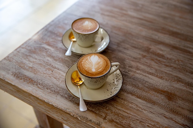 Two stylish white cups of cappuccino with latte art on saucer and golden spoons on light wooden table